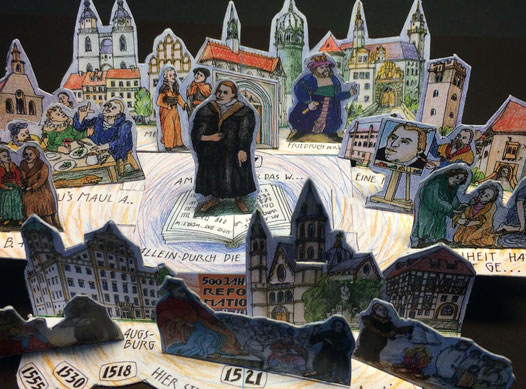 Bild mit gezeichneten Gebäuden der Stadt Wittenberg. Der Reformator Martin Luther steht in der Mitte auf einer Bibel. Alle Teile sind nach Zeichnungen geduckt und ausgestanzt. Sie stehen auf einer Streichholzschachtel, deren Boden man sehen kann. 