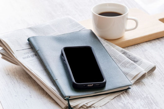 英字新聞の上に置かれた手帳とスマートフォン。カッティングボードに置かれたコーヒーのマグカップ。