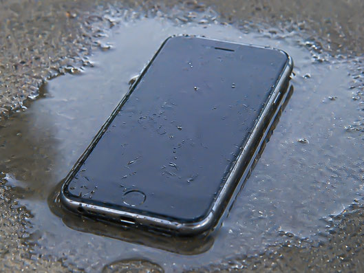 濡れた地面の上にiPhoneが置かれ、本体や画面に水滴が付着している様子を写した写真。iPhoneの水濡れや水没による不具合、水没修理の症状を連想しやすい状態。
