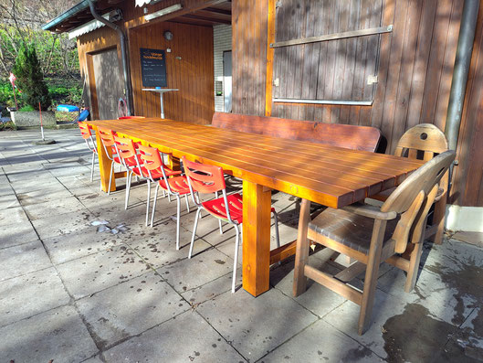 Langer, rechteckiger Holztisch mit glatter Oberfläche steht auf einer gepflasterten Terrasse vor einer Holzwand. An einer Längsseite sind mehrere rote Stühle mit Metallbeinen aufgereiht, an der gegenüberliegenden Seite zwei hölzerne Stühle mit Armlehnen.