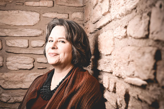 Iris Maaß blickt lächelnd vor einer Mauer stehend