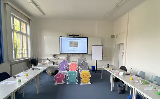 "Das Bild zeigt einen Seminarraum der VHS Hamburg. An der Vorderseite hängt ein großer Bildschirm. Auf den Tischen liegen Materialien und in der Bildmitte sieht man große Pappaufsteller."