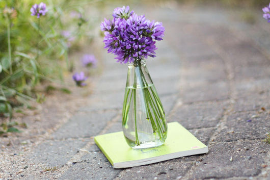 石畳の上に置かれた黄緑色の背表紙のスケッチブック。その上に紫色の花が生けられたガラスの花瓶が置かれている。