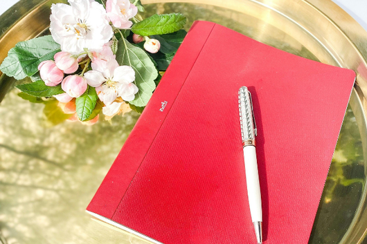 ゴールドのトレイの上に置かれた赤色の背表紙のノートとボールペン。桜の花。