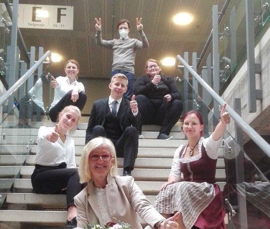 Gruppe von Absolvent:innen sitzt fröhlich auf einer Treppe, zeigt Urkunden und feiert den Lehrabschluss im Beruf Restaurantfachmann/-frau