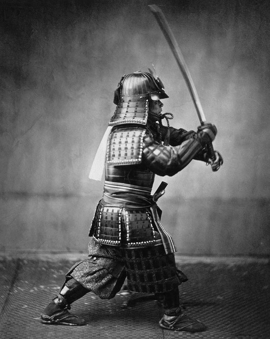A samurai in full armor raises a katana mid-strike in a dramatic pose, captured in a monochrome setting with textured backdrop.