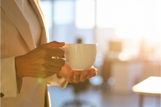 夕暮れのオフィスでコーヒーカップを両手で抱えるジャケット姿の女性。