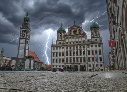 KI-bearbeitetes Foto des Augsburger Rathausplatzes bei Nacht: Blitzeinschläge über dem Perlachturm und dem historischen Rathaus mit Touristeninformation.ennen. 