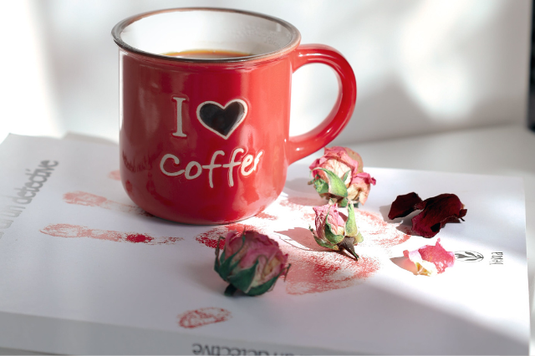 日差しが差し込むオフィスのデスク。本の上にコーヒーの入った赤のマグカップ。バラの花のポプリ。