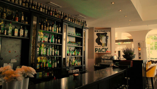 Das Ambiente im Ristorante La Volpe in Düsseldorf, auf dem klassische Poster, die beleuchtete Bar, der schwarze Tresen und eingedeckte Tische mit Tischdecke zu sehen sind.