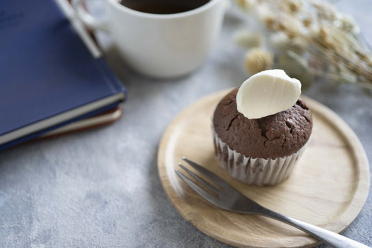 ホワイトチョコがのったマフィンのお皿。フォーク。積まれた本。コーヒーの入ったマグカップ。ドライフラワー。