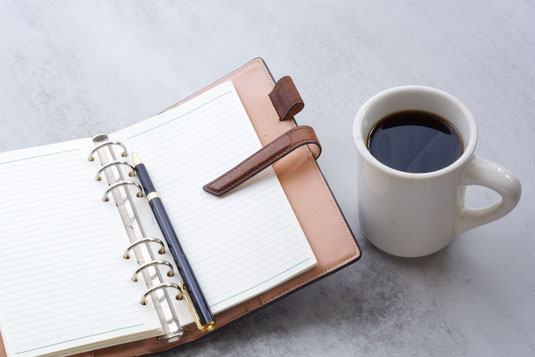 革のシステム手帳にはさまれたボールペン。コーヒーの入ったマグカップ。