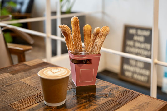カフェの店内。テーブルに置かれたカフェラテの紙カップとチュロス。営業中。