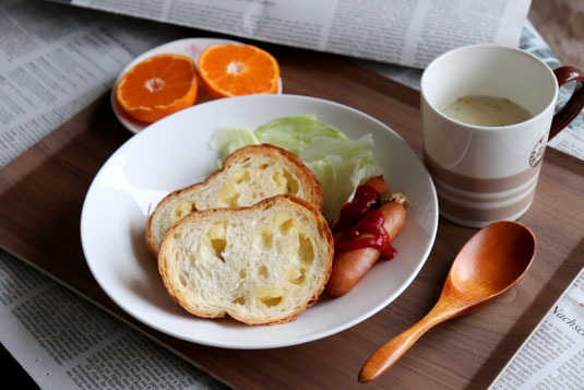 新聞の上に置かれた朝食のトレイ。チーズのパンとウインナーのお皿。半分にカットされたオレンジ。ポタージュスープのマグカップ。
