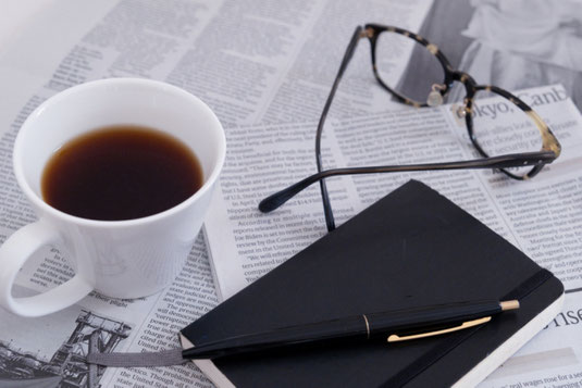 英字新聞の上に置かれたコーヒーの入ったマグカップ、手帳と万年筆、セルロイドフレームの眼鏡。