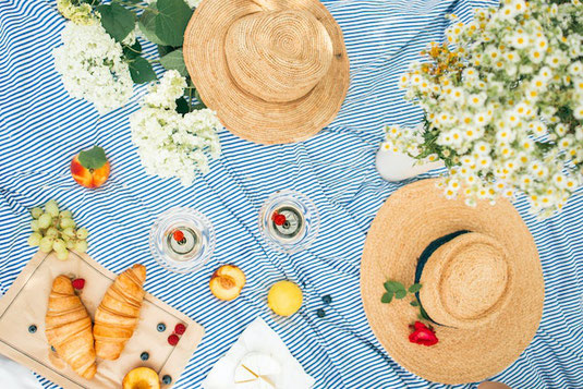 ストライプのレジャーシートのうえに麦わら帽子。フルーツ、飲み物のグラス、クロワッサンを広げてピクニック。休日のひととき。