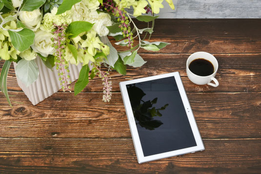 テーブルに飾られたバラのフラワーアレンジメント。隣にタブレットとコーヒーの入ったマグカップ。
