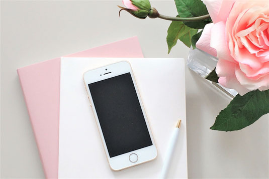 積まれたノートの上にスマートフォンとボールペンが置かれている。ガラスの一輪挿しにピンク色のバラの花が飾られている。