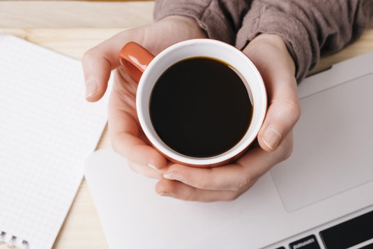 ノートパソコンと広げたリングノートの前でコーヒーのマグカップを両手で抱える。