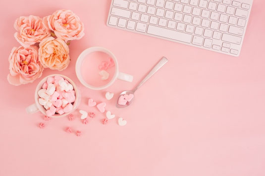 ハート形のマシュマロが詰められたマグカップがパソコンのキーボードの前に置かれている。ピンク色のバラの花。