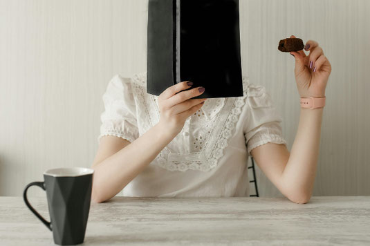 眼鏡を片手に持ち本を読む女性。紺のブラウスとカーキのパンツ姿。