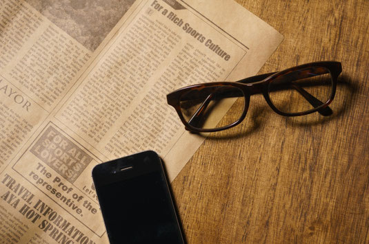 デスクに広げられた英字新聞。傍らにスマートフォンとセルロイドフレームの眼鏡。