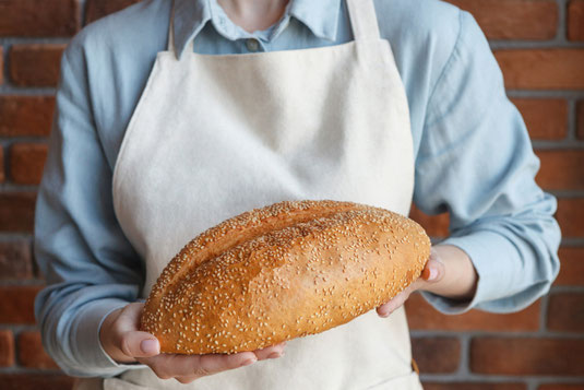 両手でゴマのフランスパンを抱えるベーカリーの店員さん。