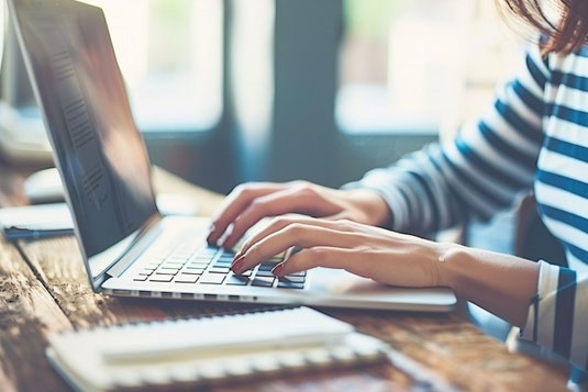 オフィスのデスクでノートパソコンに向かって作業するボーダーカットソー姿の女性。