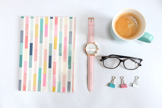 カラフルな背表紙の手帳。ピンクの革の腕時計。コーヒーの入ったモスグリーンのマグカップ。黒縁の眼鏡。3色のダブルクリップ。