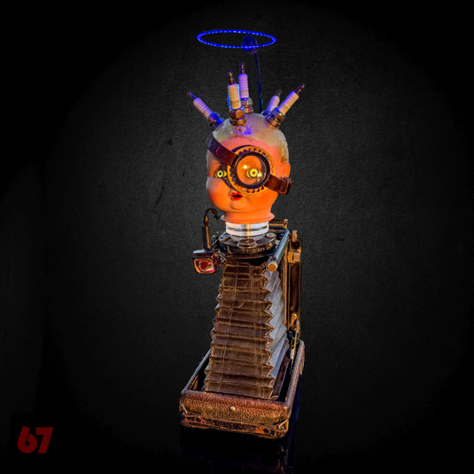 Strampelchen is an assemblage artwork made of historic parts: doll head, spark plugs, plate camera. Illuminated by LEDs and animated with an integrated motor.