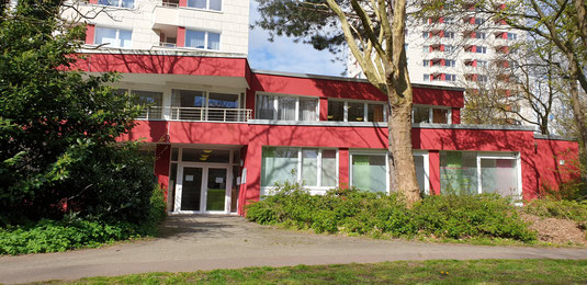 Rotes zweistöckiges Gebäude mit großen Fenstern und Eingangstür Praxis, umgeben von Bäumen und Grünflächen