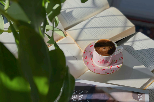 テーブルいっぱいに広げられた本。その上に置かれたコーヒーの入ったサクランボ模様のカップ＆ソーサ。観葉植物のグリーンの葉。