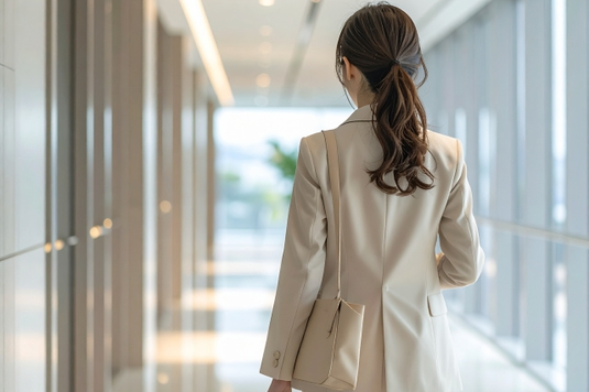 オフィスの廊下をトートバッグを肩にかけて歩くスーツ姿の女性の後ろ姿。