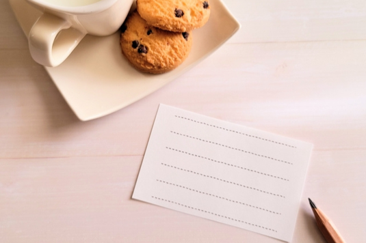 チョコチップクッキーが添えられたホットミルクのマグカップ。メモ用紙と鉛筆。
