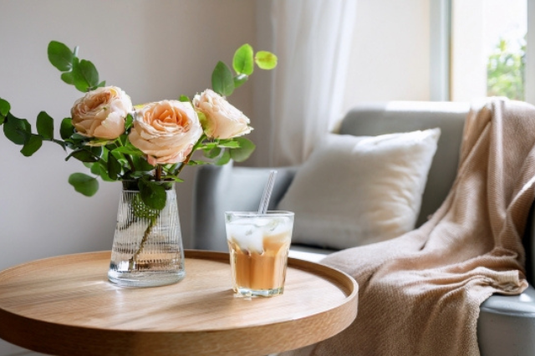 クッションとリネンストールが置かれたソファ。サイドテーブルにバラの花瓶とアイスラテのグラス。休日。