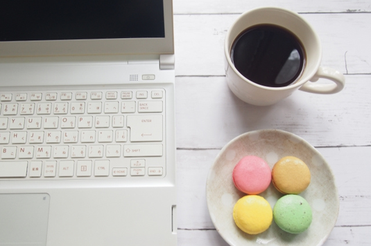 ノートパソコンのとなりにコーヒーのマグカップとパステルカラーのマカロンがのったお皿。