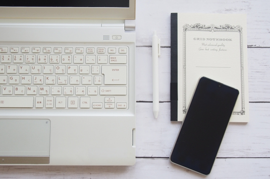 積まれたリングノートとスケジュール帳の上に置かれた白の電卓。ボールペンダブルクリップ。ボールペン。スマホ。