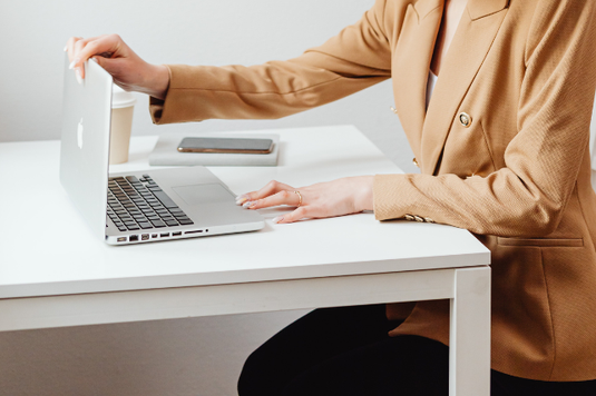 オフィスのフリースペースでノートパソコンを開く女性。ベージュ色のジャケットに黒のパンツ姿。テイクアウトコーヒー。手帳とスマホ。