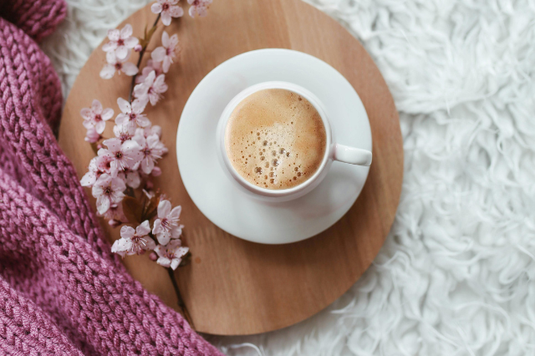 毛足の長いシャギーラグの上に置かれた木製のトレイ。コーヒーの入ったカップ＆ソーサと桜の小枝。ピンク色の毛糸のひざかけ。