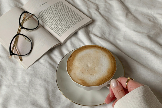 クロスが掛けられたテーブルに置かれた本。開かれたページの上に眼鏡が置かれている。カフェラテのカップに手を添えるセーター姿の女性。