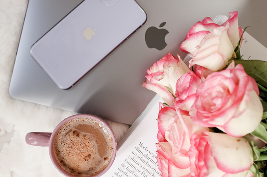 ノートパソコンとスマートフォン。ページが開かれた洋書の上に無造作に置かれたピンクのバラの花たち。カフェラテのマグカップ。