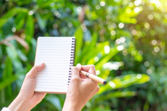 グリーンがまぶしい庭でリングメモにペンで書き込もうとする女性の指先。育児休業。