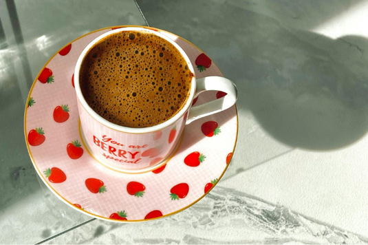 コーヒーの入ったイチゴ柄のカップ＆ソーサ。マーブル調のテーブルに日差しがさしている。