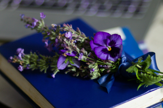 ノートパソコンの前に置かれた青色の背表紙の本。本の上に置かれたラベンダーとパンジーの花。