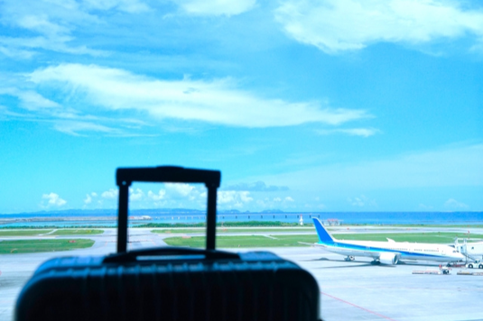 空港の窓辺に置かれたスーツケース。窓の向こうに青空と駐機する飛行機。