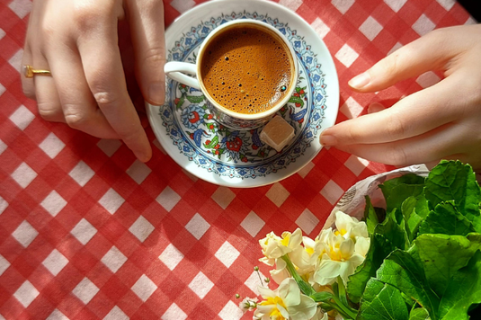 コーヒーの入ったカップ＆ソーサに両手を添える女性。フリージアの鉢植。赤のギンガムチェックのテーブルクロス。