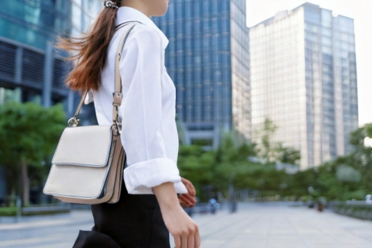 ビルが立ち並ぶオフィス街を歩く女性。白シャツに黒のタイトスカート、ショルダーバッグを下げて。