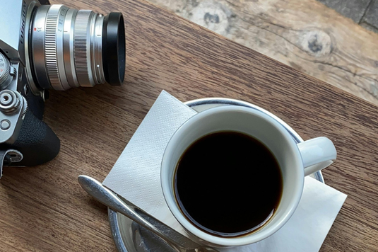 木製のテーブルに置かれたカメラとコーヒーの入ったカップ＆ソーサ。