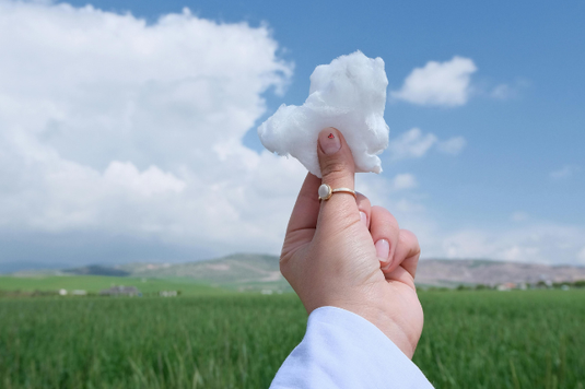 青空に浮かぶ雲に向かってハート形の綿を掲げる女性の指先。指にファッションリングをはめて。