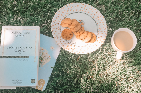 芝生の上に置かれた２冊の本。チョコチップクッキーのお皿。ミルクティーのカップ。